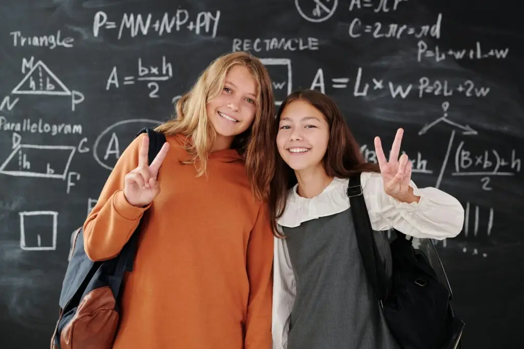 Two smiling students with backpacks in front of a math chalkboard in a classroom.