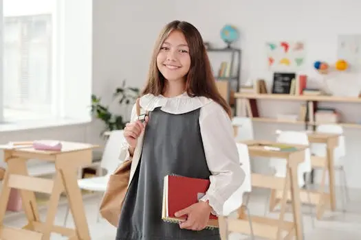 Smiling teenage girl in a school uniform holding notebooks in a bright classroom.
