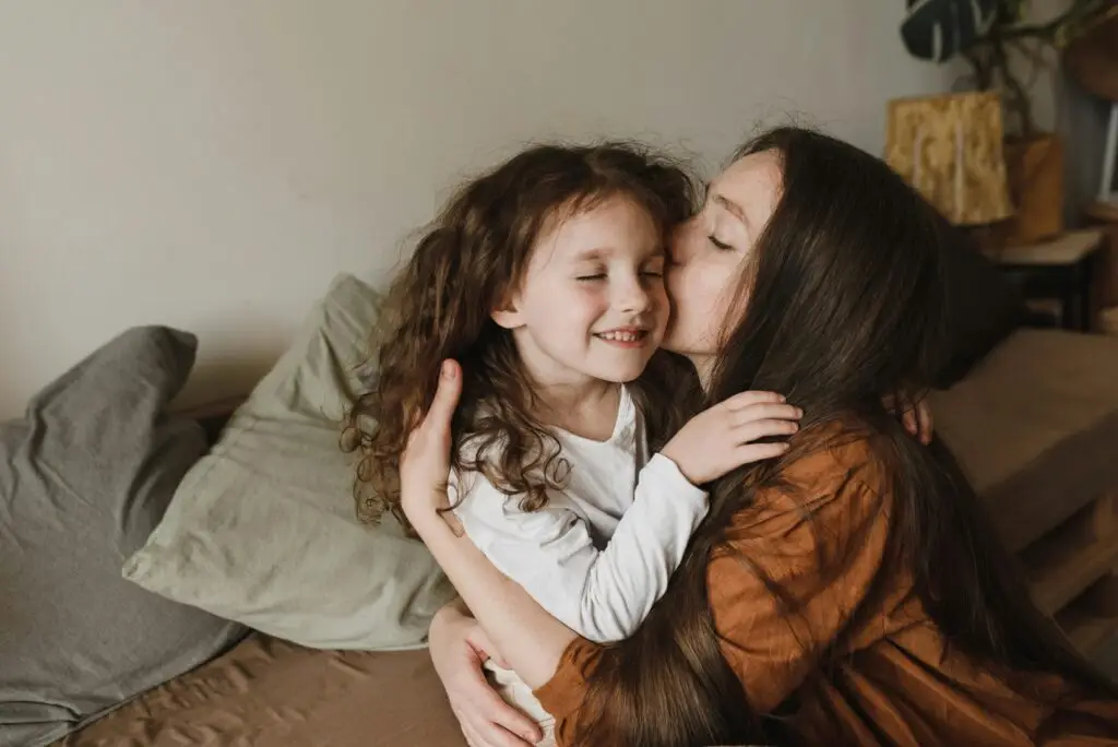 A touching moment of a mother kissing her daughter indoors, radiating love and warmth.