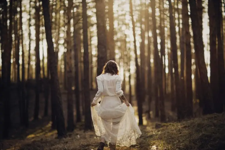 A woman in a white dress peacefully walks through a sunlit forest, capturing tranquility in nature.